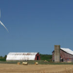 A windmill next to a barn with hay bales in front.