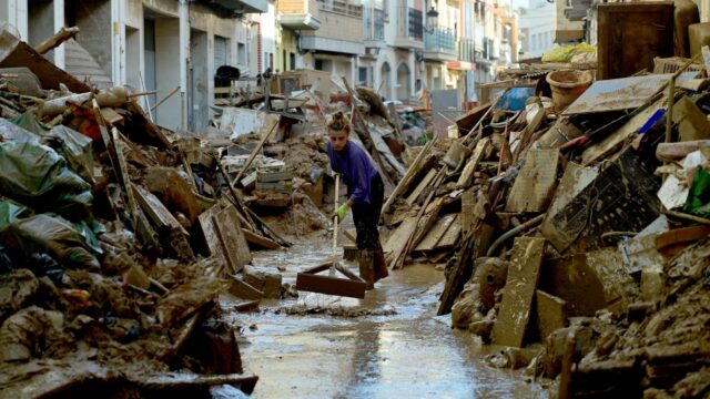 a woman in a purple shirt uses a broom to push away debris in a street full of debris on either side. she is ankle-deep in water. There is light coming from behind her. The street is narrow.