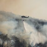 A helicopter sprays a liquid over a forest engulfed by smoke