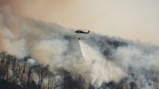 A helicopter sprays a liquid over a forest engulfed by smoke