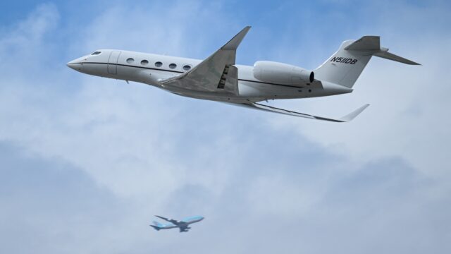 A small private jet flies through a blue sky with a passenger jet in the background