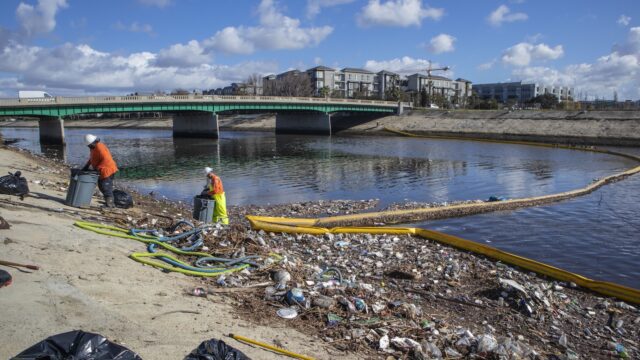 Workers on the banks of a creek collect trash using a large boom. In the distance are a bridge and tall city buildings.