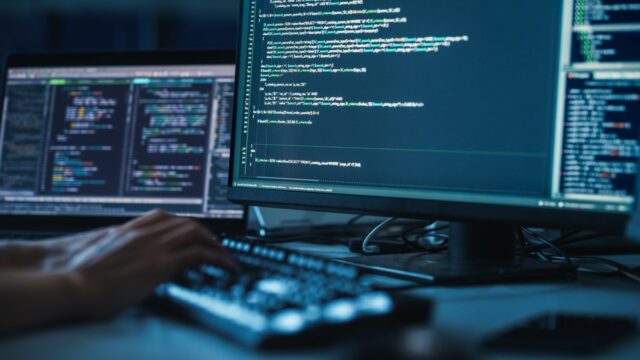 Close up of person's hands on keyboard, coding with monitors displaying code all around.