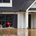 Two homeowners use buckets to bail water through the window of their flooded home.