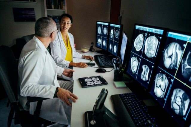 Two doctors sitting at a large computer desk talking about a patient's CT scans displayed on the monitors.