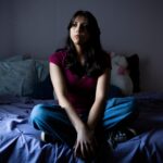 A teenage girl in a red tee shirt and jeans sits pensively on a bed