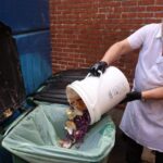 A restaurant worker empties out a bucket of food scraps into a trash can