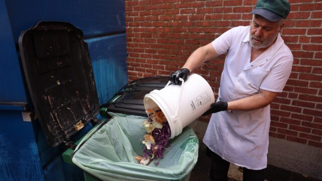 A restaurant worker empties out a bucket of food scraps into a trash can