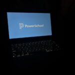 a laptop in a dark room with the laptop keyboard lit up, and the display reads PowerSchool with its corporate logo.