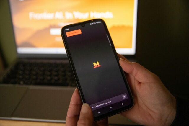 A person holds a telephone displaying the Le Chat conversational tool in front of a computer open to a page from artificial intelligence company Mistral AI